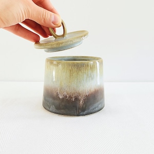Puede incluir: Un tarro de cerámica con tapa, que presenta un degradado de colores de marrón a verde. El tarro tiene forma cilíndrica con una base ligeramente más ancha. La tapa tiene un asa de bucle. El tarro probablemente se utiliza para el almacenamiento.