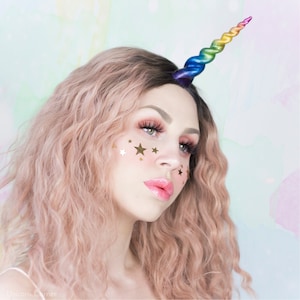May include: A woman with pink curly hair wearing a rainbow unicorn horn headband with gold star shaped stickers on her face.