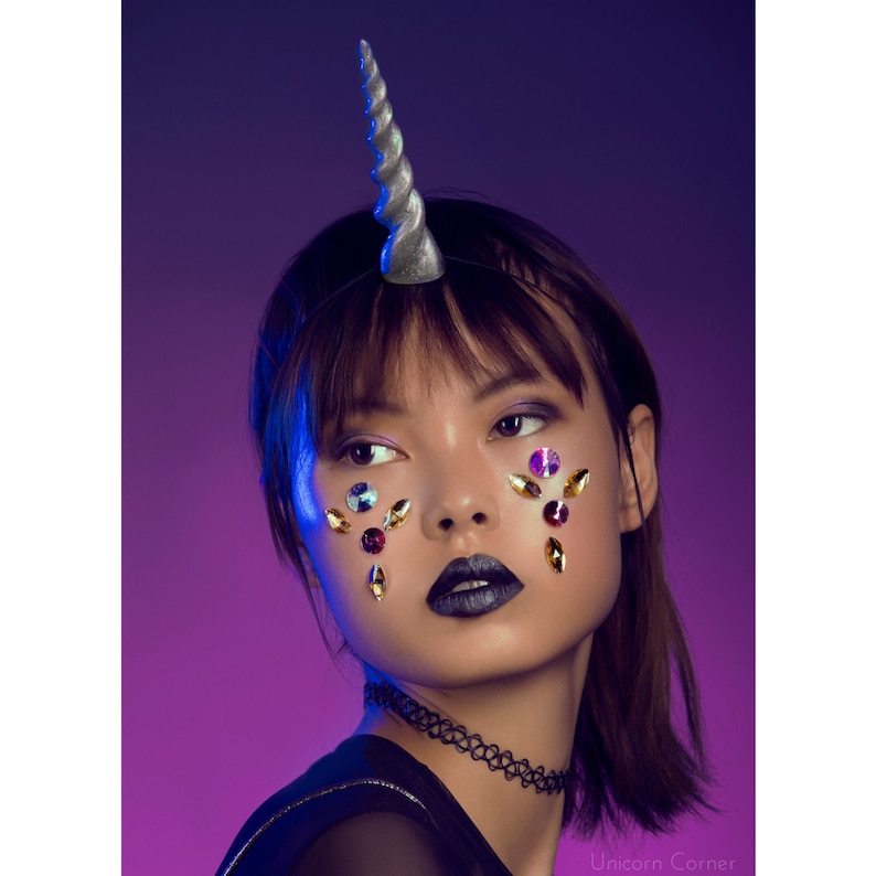 May include: A woman with a silver unicorn horn headband and colorful rhinestones on her face. She is wearing a black choker necklace and a black shirt. The background is a vibrant purple.