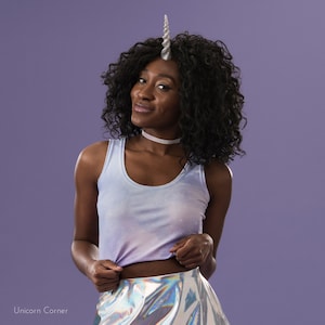 May include: A woman wearing a light blue tank top with a silver unicorn horn headband and a silver iridescent skirt. She is standing in front of a purple background.