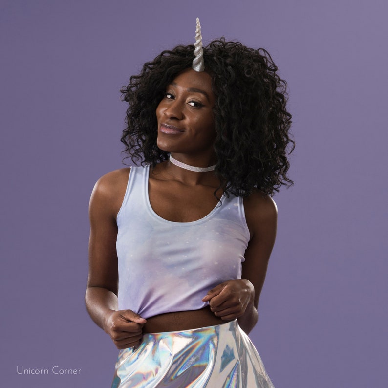 May include: A woman wearing a light blue tank top with a silver unicorn horn headband and a silver iridescent skirt. She is standing in front of a purple background.