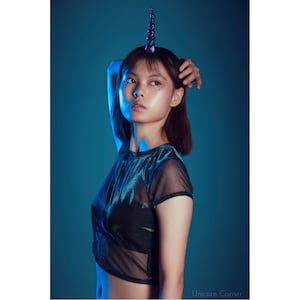 May include: A woman with short brown hair is wearing a black mesh top and a purple unicorn horn headband. She has blue eyeshadow and glitter on her face. The background is a solid blue.