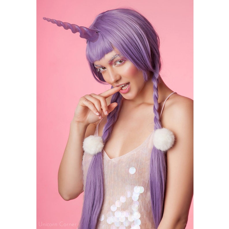 May include: A woman with long, purple braids and a unicorn horn headband is wearing a sparkly, light pink dress. She is biting her fingernail and looking at the camera.