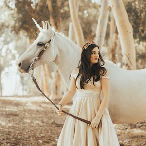 May include: A woman in a long, off-white dress with a gold crown holds the reins of a white unicorn with a horn. The unicorn is standing in a forest setting.