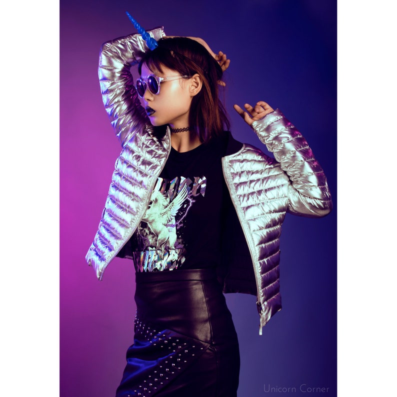 May include: A woman wearing a silver puffer jacket with a black t-shirt featuring a unicorn graphic, black leather skirt with silver studs, and blue unicorn horn headband. The background is a gradient of purple and blue.
