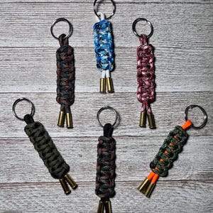 May include: A collection of six paracord keychains in various colors, including brown, blue, red, green, black, and orange. Each keychain features a braided design and is accented with brass bullet casings. The keychains are arranged on a wooden surface.