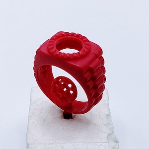 May include: A red wax ring with a circular design in the center. The ring is on a white foam stand.