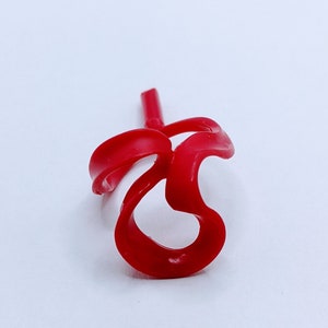 Puede incluir: Un objeto de plástico rojo brillante y abstracto con un diseño curvo y fluido. El objeto tiene una protuberancia en forma de tallo y una forma de bucle, posiblemente un elemento decorativo o un componente. El fondo es blanco liso, resaltando el color vibrante y la forma única.