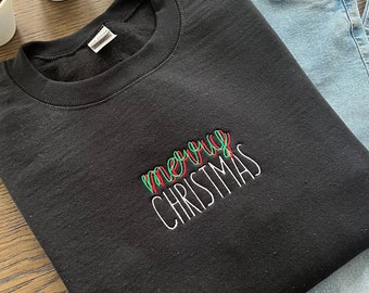 Könnte beinhalten: Schwarzes Sweatshirt mit Rundhalsausschnitt und dem gestickten Text „merry CHRISTMAS“ in Rot, Grün und Weiß. Das Sweatshirt wird mit hellblauen Jeans kombiniert. Zwei kleine Topfpflanzen befinden sich im Hintergrund.