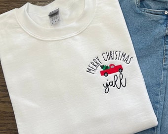 Könnte beinhalten: Weißer Pullover mit dem Text "Merry Christmas Y'all" und einem roten LKW mit einem Weihnachtsbaum in der Ladefläche. Der Pullover liegt auf einer blauen Jeans.
