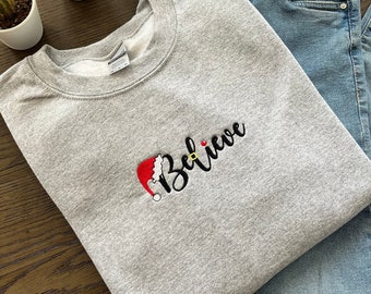 Könnte beinhalten: Ein grauer Pullover mit dem Wort "Believe" in Schwarz gestickt, darüber ein roter Weihnachtsmann-Hut.