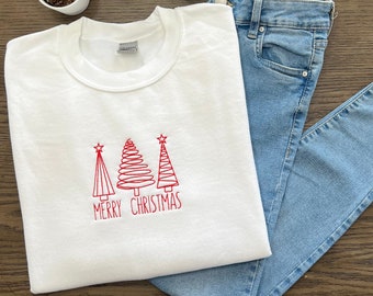 Könnte beinhalten: Weißer Pullover mit roten gestickten Weihnachtsbäumen und dem Text "Merry Christmas". Der Pullover ist gefaltet und liegt auf einer hellblauen Jeans.