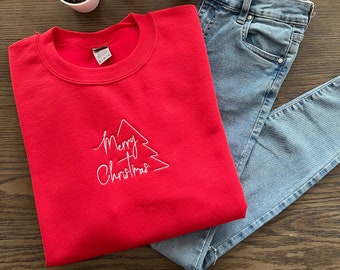 Könnte beinhalten: Roter Pullover mit einem weißen gestickten Weihnachtsbaum und dem Text "Merry Christmas". Der Pullover ist gefaltet und liegt auf einer hellblauen Jeans.
