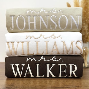 May include: Three stacked towels with embroidered text. The top towel is beige with white text that reads "mrs. JOHNSON". The middle towel is white with gold text that reads "mrs. WILLIAMS". The bottom towel is brown with white text that reads "mrs. WALKER".