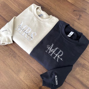 May include: Two sweatshirts, one beige and one black, with the words "MRS Williams" and "MR Williams" embroidered on them. The beige sweatshirt has the date "10.01.2023" on the sleeve, and the black sweatshirt has the date "10.20.2023" on the sleeve.
