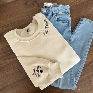 K&ouml;nnte beinhalten: Ein beiger Pullover mit dem Text "Cat Mom" auf der Schulter und "Louie Smootkey" mit einem Pfotenabdruck auf dem &Auml;rmel gestickt.