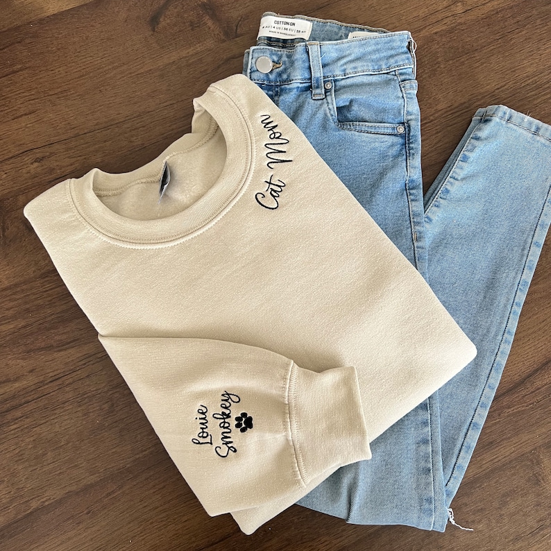 K&ouml;nnte beinhalten: Ein beiger Pullover mit dem Text "Cat Mom" auf der Schulter und "Louie Smootkey" mit einem Pfotenabdruck auf dem &Auml;rmel gestickt.