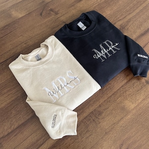 May include: Two sweatshirts, one beige and one black, with embroidered text. The beige sweatshirt says "MRS Williams" and the black sweatshirt says "MR Williams". Both sweatshirts have the date "10.20.2023" embroidered on the sleeve.