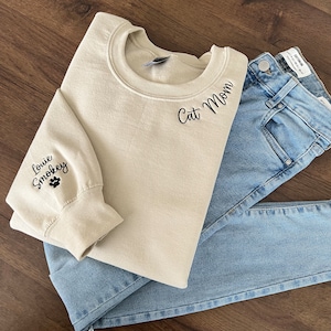 K&ouml;nnte beinhalten: Ein beiger Pullover mit dem Schriftzug "Cat Mom" auf der Vorderseite und "Louie Smokey" mit einem Pfotenabdruck auf dem linken &Auml;rmel bestickt.