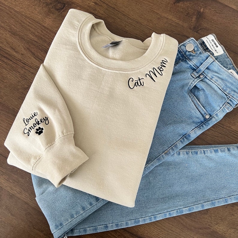 K&ouml;nnte beinhalten: Ein beiger Pullover mit dem Schriftzug "Cat Mom" auf der Vorderseite und "Louie Smokey" mit einem Pfotenabdruck auf dem linken &Auml;rmel bestickt.