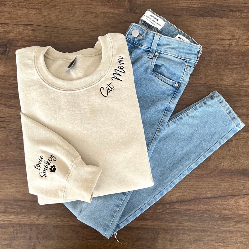 K&ouml;nnte beinhalten: Ein beiger Pullover mit dem Text "Cat Mom" auf dem Ausschnitt und "Louie Smokley" sowie einem gestickten Pfotenabdruck auf dem linken &Auml;rmel.