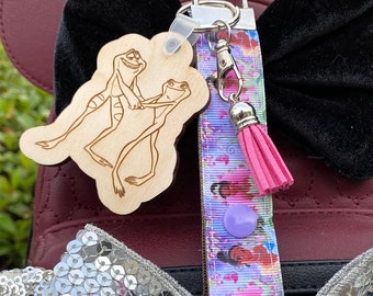 Può includere: Un portachiavi in legno con un design tagliato al laser di due rane che ballano. Il portachiavi è attaccato a un cordino fantasia con un nappa rosa e un bottone viola. Lo sfondo include uno zaino bordeaux e fiocchi di velluto nero.