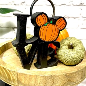 May include: Black wooden "LOVE" sign with a pumpkin Mickey Mouse head in the center. The sign is on a tiered wooden tray with two small pumpkins.
