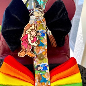May include: A burgundy backpack with black sequin accents and a rainbow bow. A black velvet bow and a keychain with a cartoon character charm and a wristlet strap featuring cartoon characters are also visible.