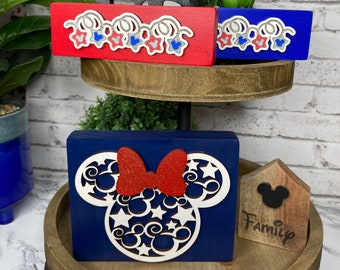 Peut inclure: Un plateau en bois à étages avec trois décorations en bois rouge, blanc et bleu. La décoration du haut est une silhouette noire de Mickey Mouse avec le mot "Love" devant. La décoration du milieu est une guirlande rouge, blanche et bleue avec des têtes de Mickey Mouse. La décoration du bas est un carré bleu avec une silhouette blanche de la tête de Mickey Mouse avec un nœud rouge et des étoiles blanches. Le niveau inférieur du plateau a également une petite maison en bois avec une tête de Mickey Mouse et le mot "Family" écrit dessus.
