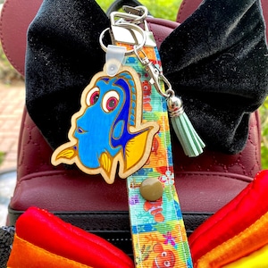 May include: A keychain featuring a wooden Dory character from Finding Nemo. The keychain is attached to a colorful lanyard with a cartoon fish pattern and a small green tassel. The background includes a burgundy backpack and a rainbow-colored bow.