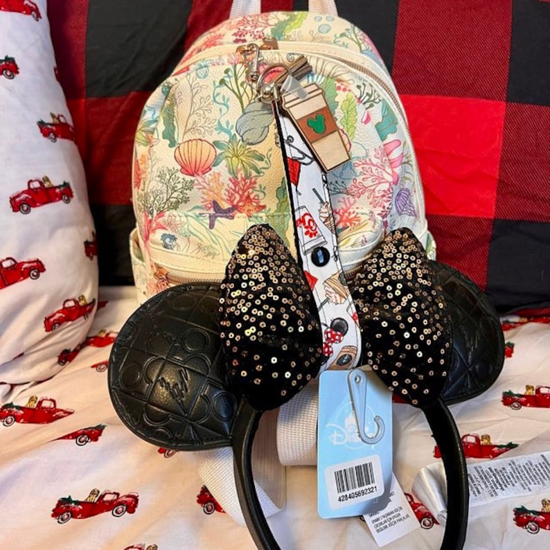 Pode incluir: Uma mochila branca com estampa floral com um chaveiro combinando, orelhas da Minnie Mouse com lantejoulas pretas e uma faixa de cabe&ccedil;a preta. As orelhas t&ecirc;m um design acolchoado. Uma etiqueta com um c&oacute;digo de barras est&aacute; presa na faixa de cabe&ccedil;a.