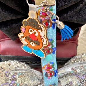 Pode incluir: Um chaveiro de madeira do Mr. Potato Head com uma borla azul e um cord&atilde;o azul claro. O chaveiro apresenta um Mr. Potato Head castanho com chap&eacute;u preto, bigode e nariz vermelho. O cord&atilde;o tem um padr&atilde;o repetido de personagens do Mr. Potato Head.