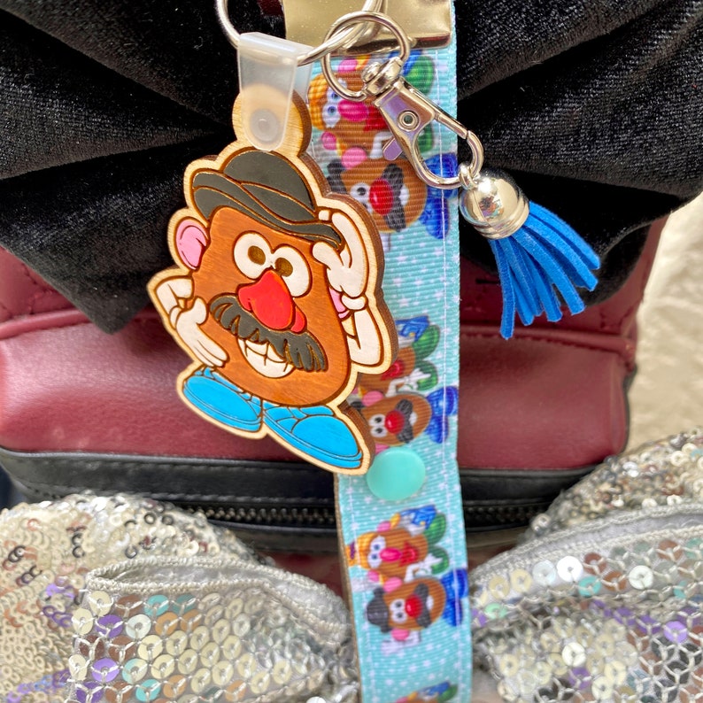 Pode incluir: Um chaveiro de madeira do Mr. Potato Head com uma borla azul e um cord&atilde;o azul claro. O chaveiro apresenta um Mr. Potato Head castanho com chap&eacute;u preto, bigode e nariz vermelho. O cord&atilde;o tem um padr&atilde;o repetido de personagens do Mr. Potato Head.
