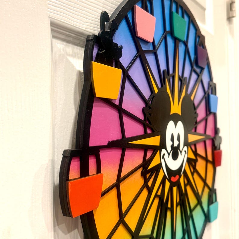 Puede incluir: Una noria de madera colorida con la cara de Mickey Mouse en el centro. La noria tiene un fondo degradado arco&iacute;ris y radios negros. Hay ocho g&oacute;ndolas coloridas alrededor de la noria.
