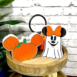 May include: A two-tiered wooden tiered tray with a white pumpkin with a green stem and a white ghost with a black bow and a black face. The ghost is wearing a red bow.