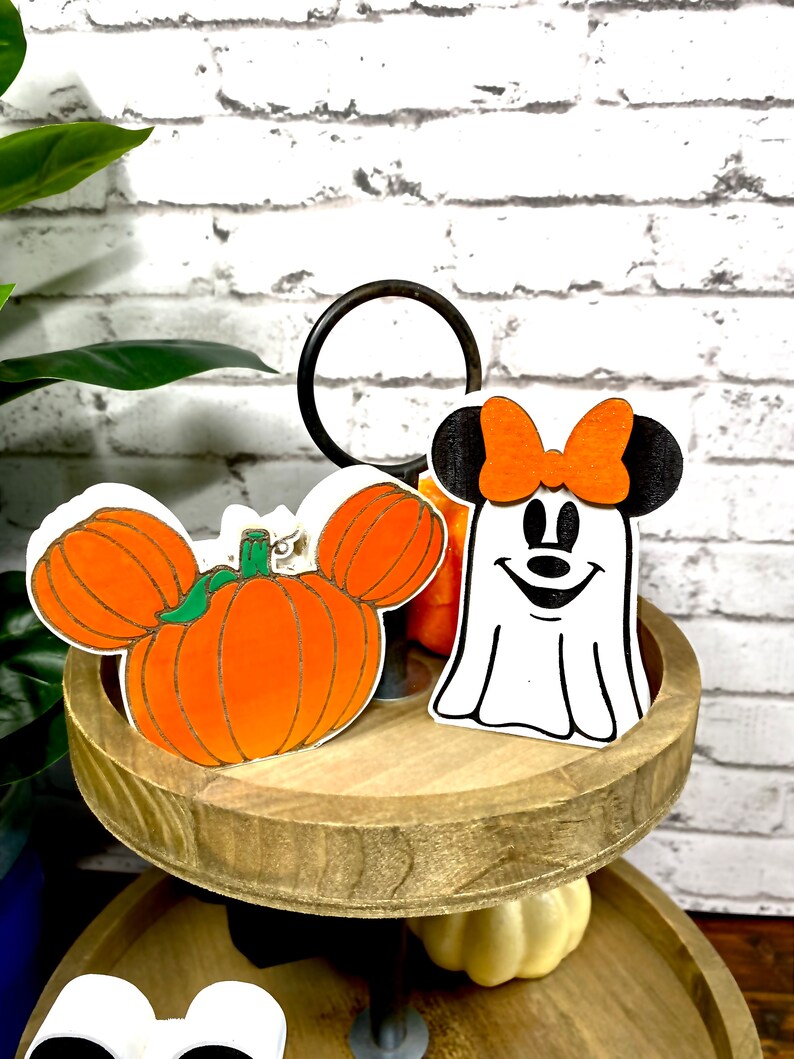 May include: A two-tiered wooden tiered tray with a white pumpkin with a green stem and a white ghost with a black bow and a black face. The ghost is wearing a red bow.