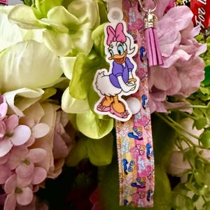 May include: A Daisy Duck keychain with a pink bow, purple dress, and orange shoes. The keychain has a pink and blue patterned strap with a silver clasp and a pink tassel. The background features pink and green flowers.