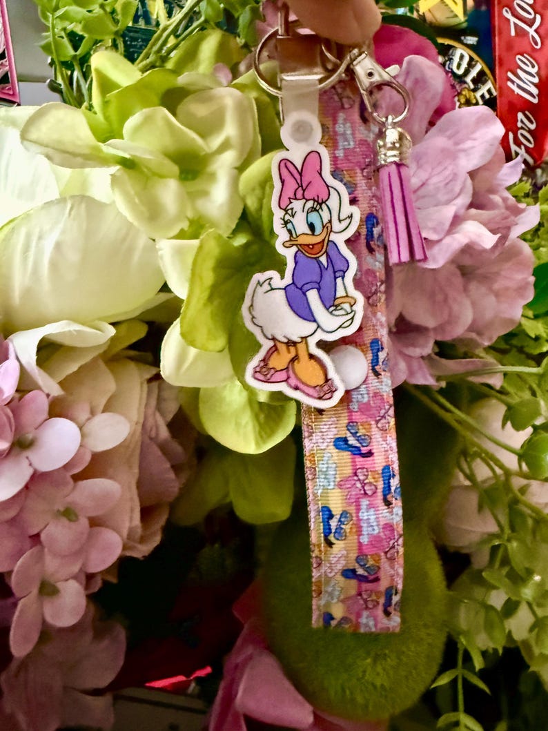 May include: A Daisy Duck keychain with a pink bow, purple dress, and orange shoes. The keychain has a pink and blue patterned strap with a silver clasp and a pink tassel. The background features pink and green flowers.