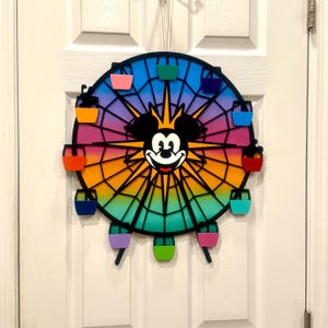 Puede incluir: Una noria de madera colorida con la cara de Mickey Mouse en el centro. La noria tiene un fondo degradado arco&iacute;ris y radios negros. Hay 10 g&oacute;ndolas coloridas alrededor de la noria.