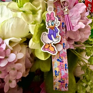 May include: A keychain featuring a Daisy Duck charm, a pink and white patterned strap, and a pink tassel. The Daisy Duck charm is wearing a purple dress and pink shoes. The strap has a pattern of butterflies and sandals.