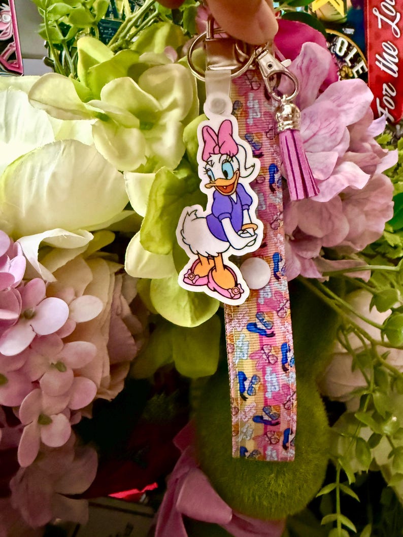 May include: A keychain featuring a Daisy Duck charm, a pink and white patterned strap, and a pink tassel. The Daisy Duck charm is wearing a purple dress and pink shoes. The strap has a pattern of butterflies and sandals.