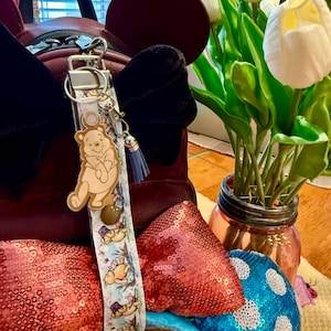 May include: A Winnie the Pooh themed keyring with a light brown bear charm and a blue tassel. The keyring strap features a light blue background with Winnie the Pooh illustrations. The background includes a maroon bag and a vase of white flowers.