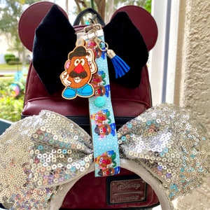 Pode incluir: Um chaveiro do Mr. Potato Head com um cord&atilde;o estampado azul e branco, um borla azul e um bot&atilde;o azul claro. O chaveiro est&aacute; preso a uma mochila bord&ocirc; com orelhas de veludo preto e um la&ccedil;o de lantejoulas.