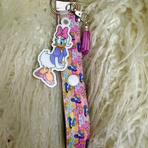 May include: A pink and yellow wristlet keychain with a Daisy Duck charm and a purple tassel. The wristlet strap features a pattern of ballet slippers and bows. The keychain has a silver-colored metal clasp and key ring.