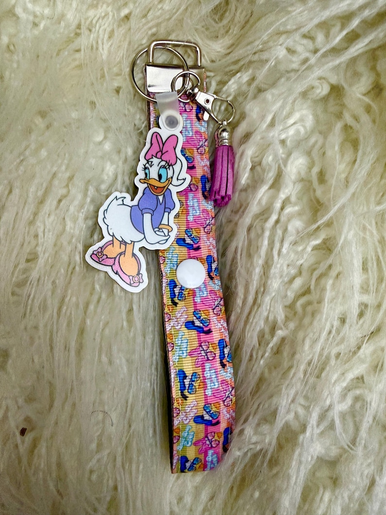 May include: A pink and yellow wristlet keychain with a Daisy Duck charm and a purple tassel. The wristlet strap features a pattern of ballet slippers and bows. The keychain has a silver-colored metal clasp and key ring.