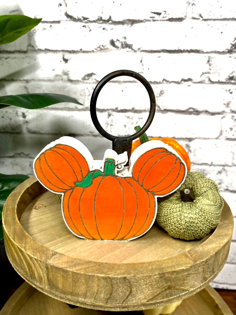 May include: A wooden tiered tray with a pumpkin shaped like Mickey Mouse's head. The pumpkin is orange with green leaves and a brown stem. The tray is brown and has a rustic look.