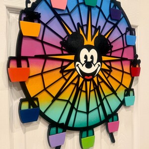 Puede incluir: Una noria de madera colorida con Mickey Mouse en el centro. La noria tiene ocho g&oacute;ndolas, cada una pintada de un color diferente: rojo, naranja, amarillo, verde, azul, morado, rosa y negro.
