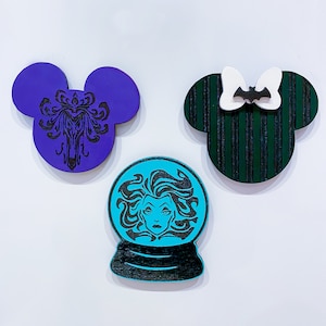 Op de afbeelding: Drie houten ornamenten in de vorm van Mickey Mouse-oren. E&eacute;n is paars met een zwart silhouet van een spookhuis, &eacute;&eacute;n is groen met zwarte strepen en een witte strik met een zwarte vleermuis en &eacute;&eacute;n is blauw met een zwart silhouet van een vrouw met lang haar.