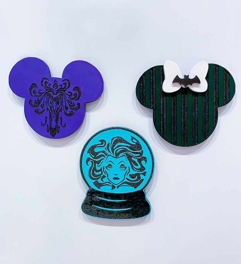 Op de afbeelding: Drie houten ornamenten in de vorm van Mickey Mouse-oren. E&eacute;n is paars met een zwart silhouet van een spookhuis, &eacute;&eacute;n is groen met zwarte strepen en een witte strik met een zwarte vleermuis en &eacute;&eacute;n is blauw met een zwart silhouet van een vrouw met lang haar.