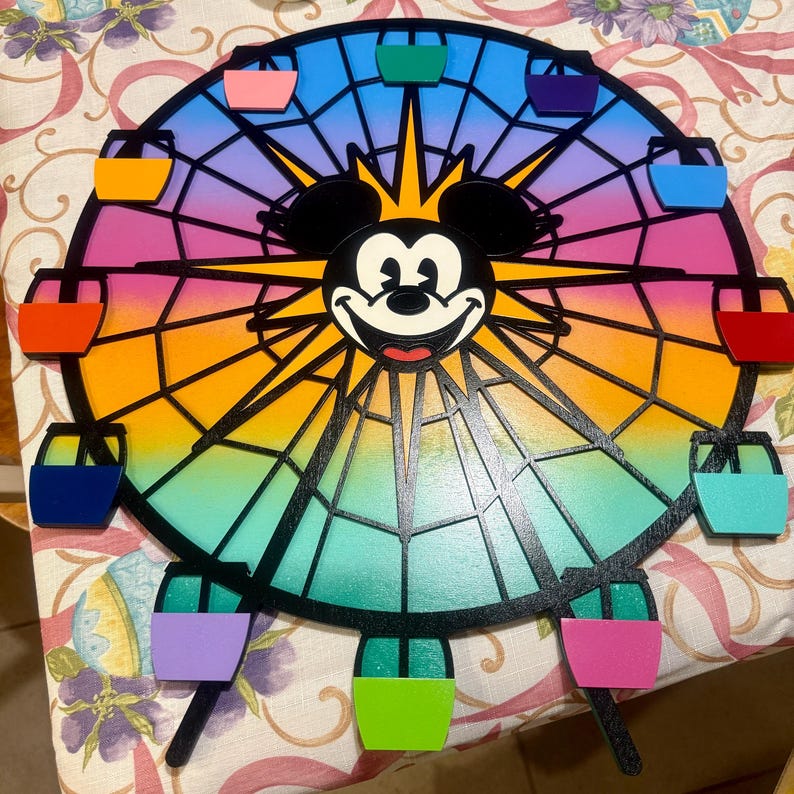 Puede incluir: Una noria de madera colorida con la cara de Mickey Mouse en el centro. La noria tiene un fondo degradado arco&iacute;ris y radios negros. Hay 10 g&oacute;ndolas coloridas alrededor de la noria.